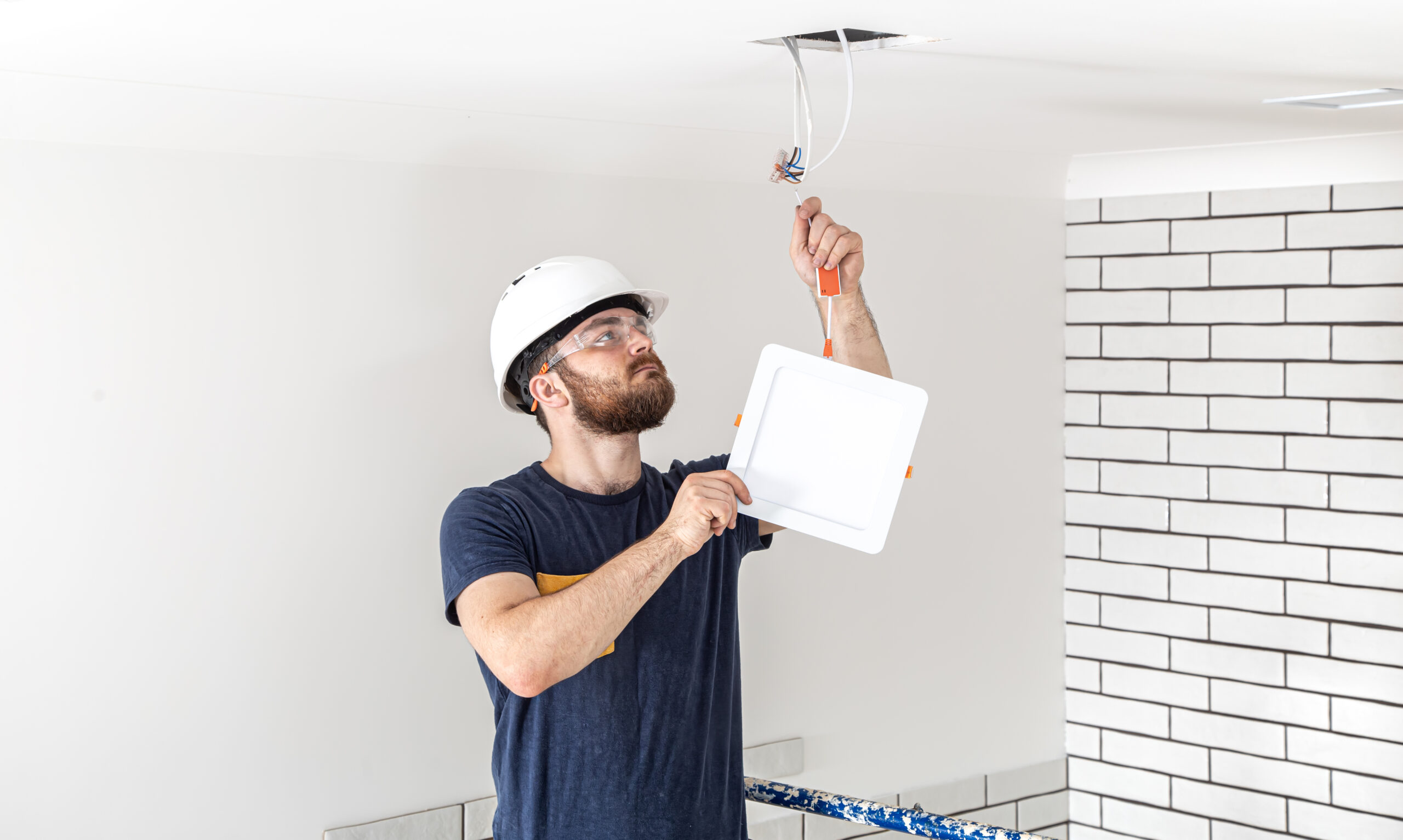 Electrician Builder with beard worker in a white helmet at work, installation of lamps at height. Professional in overalls with a drill on the background of the repair site.