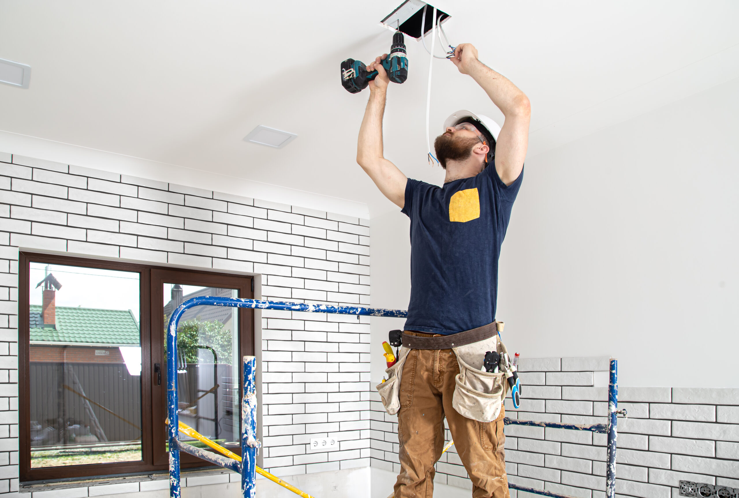 Electrician Builder at work, installation of lamps at height. Professional in overalls with a drill on the background of the repair site.